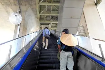 Naiki eskalator untuk ke Terminal Bus Maiko, yang terletak di kaki Jembatan Besar Selat Akashi. Itu langsung terhubung ke Stasiun JR Maiko. Pria yang lehernya menggantung topi bambu pasti adalah seorang peziarah yang menuju Tokushima di Shikoku