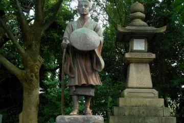 <p>Statue of Matsuo Basho, a famous poet in the Edo Period, standing in the corner of the shrine grounds with a stone lantern and tall trees</p>