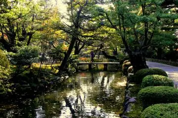<p>Trees reflected on a pond as a couple walks over one of the garden's small bridges</p>