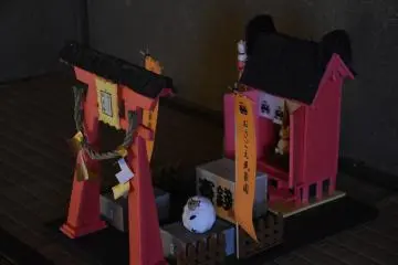 <p>Miniature shrine and torii gate displayed inside a house. The orange banner says 'Osagoe Folk Museum Village'.</p>